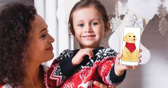 A woman and a young girl smile as the girl holds up a smartphone displaying a golden retriever wearing a bandana.