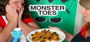 Two people present a plate of food in front of a "MONSTER TOES" banner.
