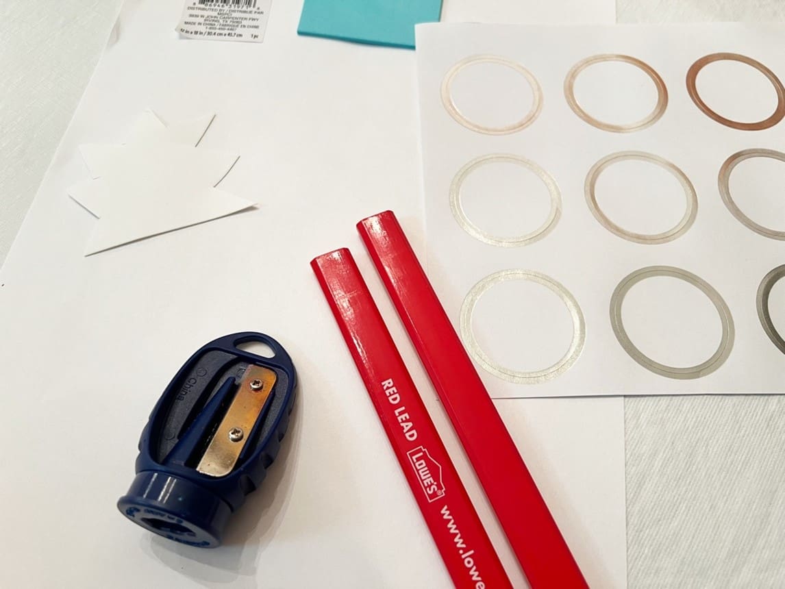 Two red pencils, a pencil sharpener, and a sheet of circular stickers on a white surface.