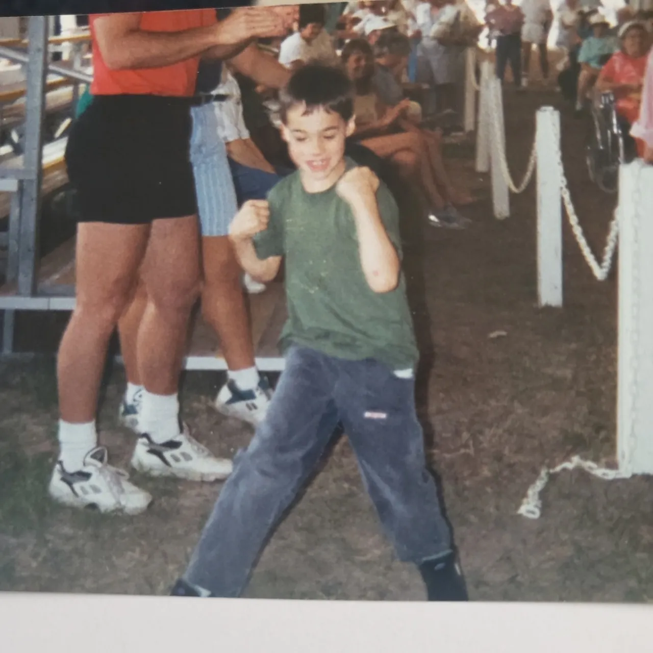 A young boy with his fists raised stands in a grassy area with people in the background.