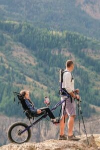 Man with a Huckleberry Hiking tandem carrier with a child in it on a rocky outcrop.