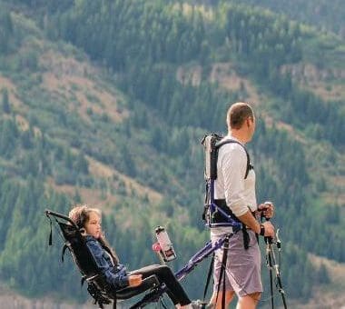 Man with a Huckleberry Hiking tandem carrier with a child in it on a rocky outcrop.
