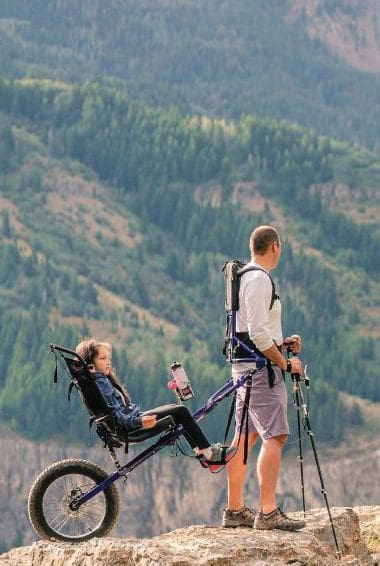 Man with a Huckleberry Hiking tandem carrier with a child in it on a rocky outcrop.
