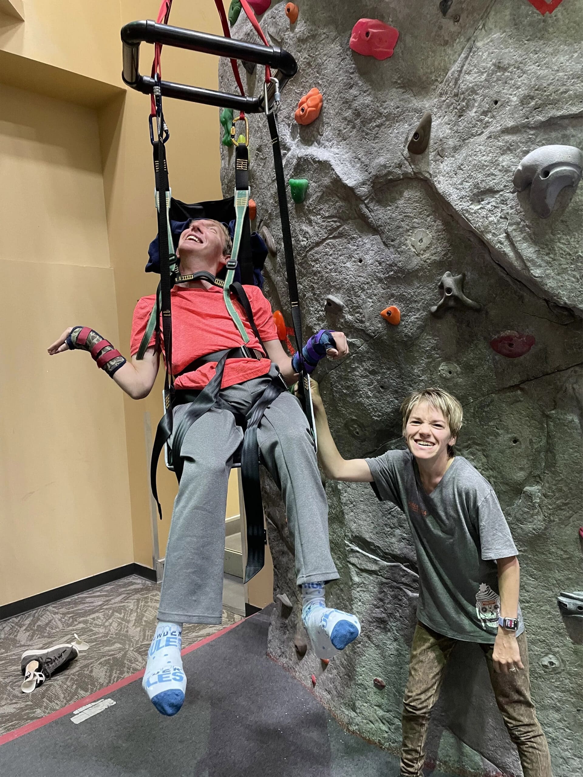 Two individuals are participating in adaptive climbing, with one person suspended in a harness and another standing next to a climbing wall.