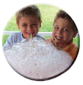 Two boys blow bubbles with straws into a large bowl of soapy water.