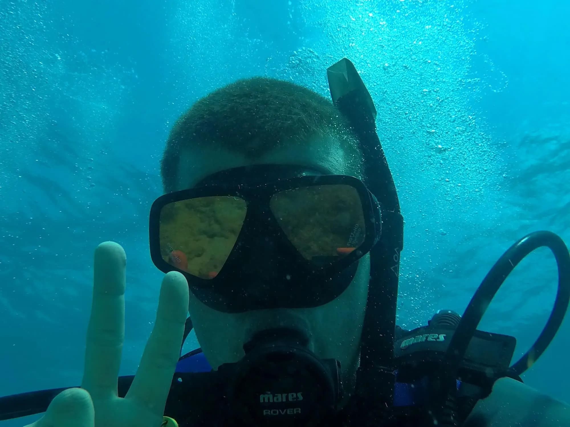 Clayton in a diving mask and scuba gear gives a peace sign underwater.