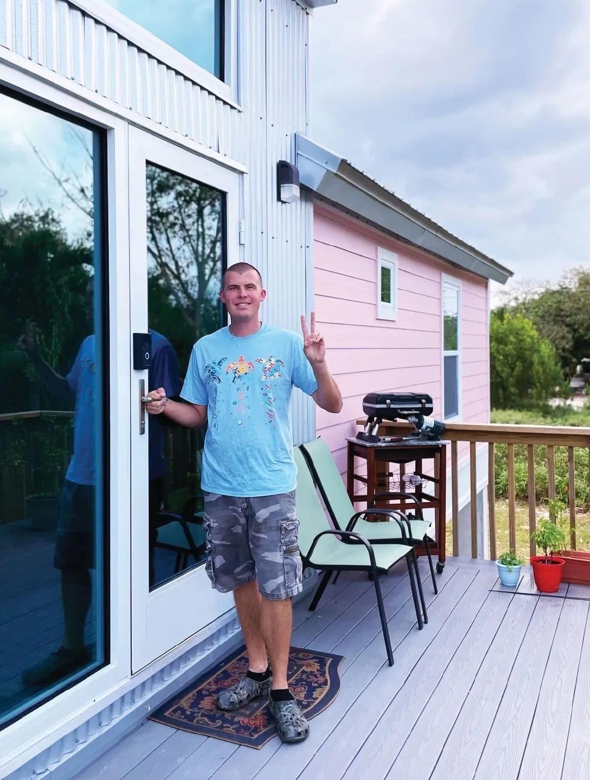 Clayton stands on a porch in front of a pink house, holding up two fingers.