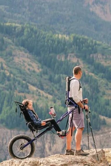 A man with trekking poles stands next to a Huckleberry Hiking device carrying a child on a mountain trail.
