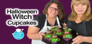 Two women hold Halloween witch cupcakes with purple and green frosting and black witch hats.