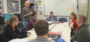 A teacher stands and speaks to a group of students seated around a table.