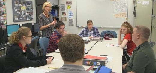 A teacher stands and speaks to a group of students seated around a table.
