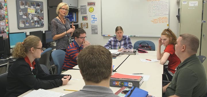 A teacher stands and speaks to a group of students seated around a table.