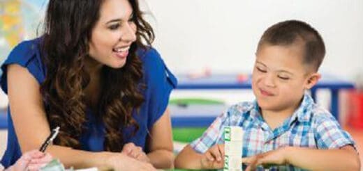 A woman in a blue shirt smiles at a boy with Down syndrome wearing a plaid shirt as they sit at a table.