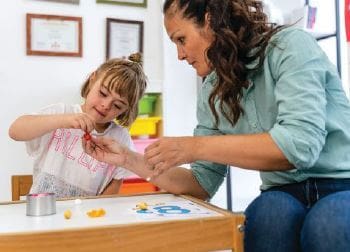 Woman helps a young child with an activity at a table.