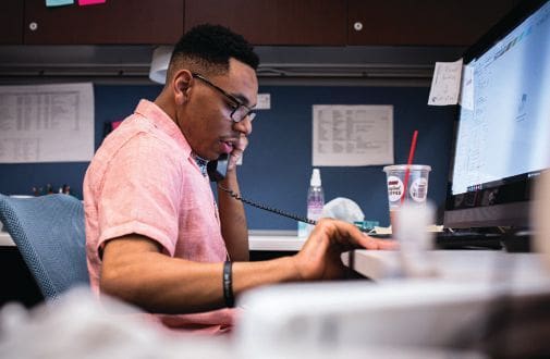 Man wearing glasses and a pink shirt talking on a landline phone while looking at a computer screen.