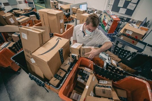 A man sorts Amazon packages in a warehouse surrounded by bins and boxes.