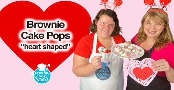 Two women hold heart-shaped brownie cake pops and a decorative heart cutout.