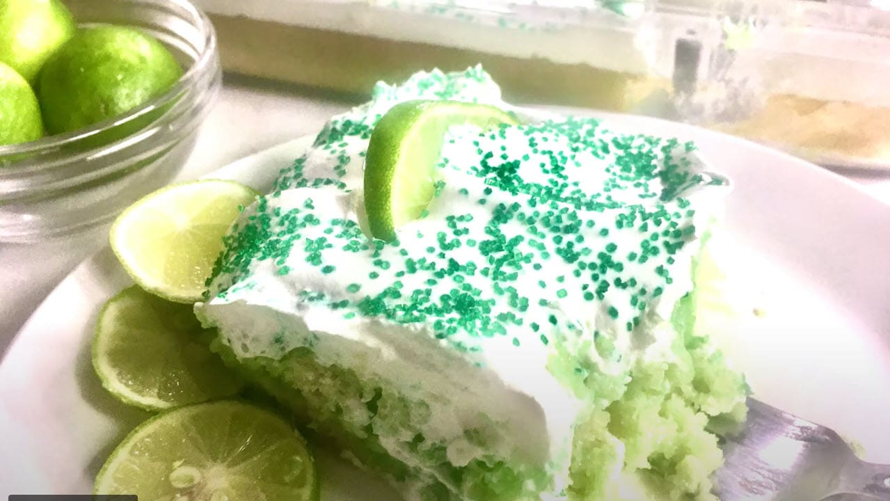 A slice of green poke cake topped with whipped cream, green sprinkles, and lime slices.