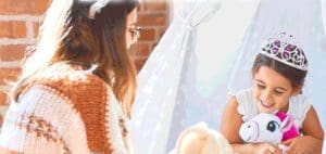 A young girl wearing a tiara smiles while holding a stuffed unicorn inside a tent.
