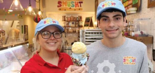 Two employees wear light blue caps and smile in front of an ice cream shop display.