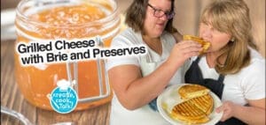 Two women prepare a grilled cheese sandwich with brie and preserves.