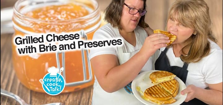 Two women prepare a grilled cheese sandwich with brie and preserves.