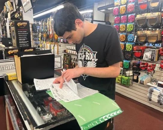Young man in a black t-shirt reviews papers at a counter in a store.