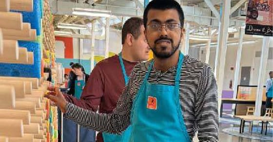 A man wearing glasses and a teal apron looks towards the camera.