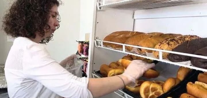 Baker in white shirt and gloves arranges loaves of bread on shelves.