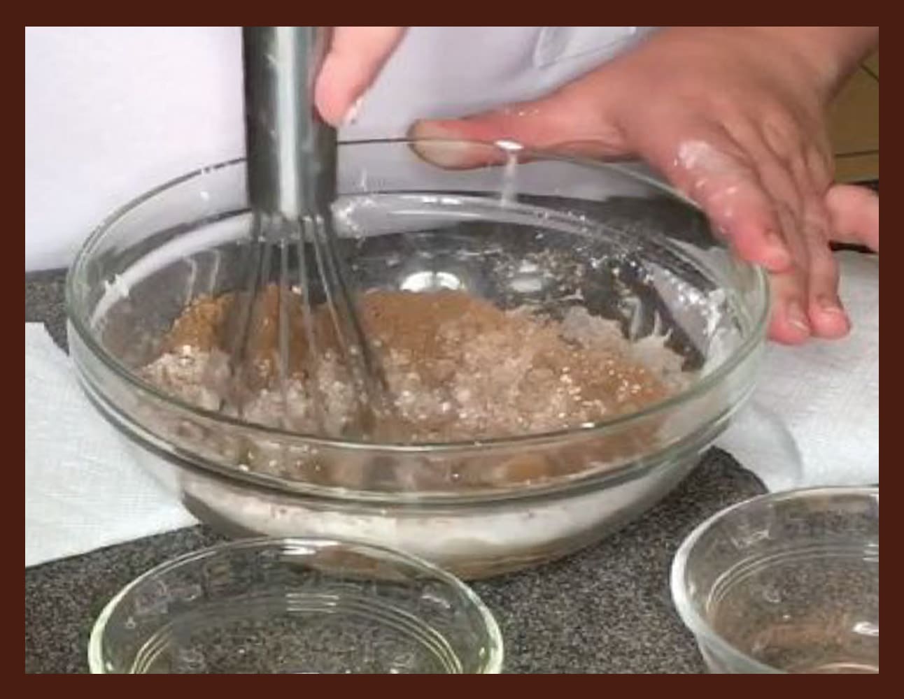 Hands whisking ingredients in a glass bowl with smaller bowls nearby.