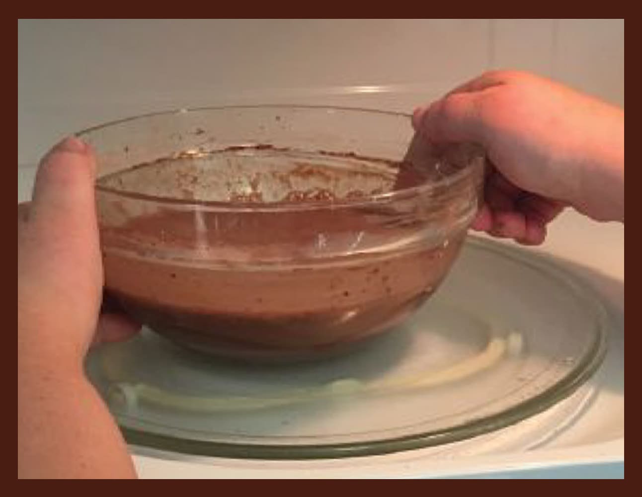 Hands mixing chocolate pudding ingredients in a glass bowl on a plate.