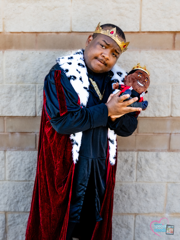 Darius wears a crown and a royal cape while holding a stuffed doll also wearing a crown.