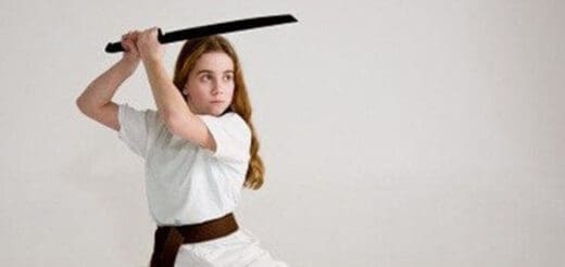 A young girl in a white gi and brown belt holds a sword above her head.