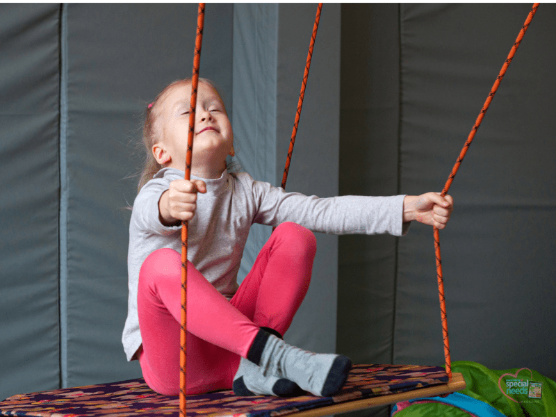 A young girl in a grey long-sleeved shirt and pink leggings sits on a swing, holding the ropes with her eyes closed.