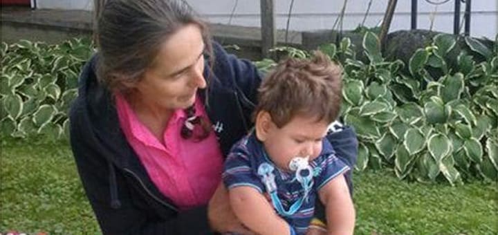 A woman holds a baby with a pacifier, with plants in the background.