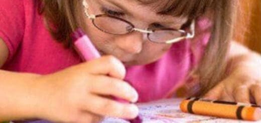 Child wearing glasses and a pink shirt draws with a pink marker and an orange crayon.