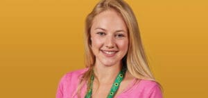 Woman with blonde hair wearing a pink sweater and a sunflower lanyard smiles.