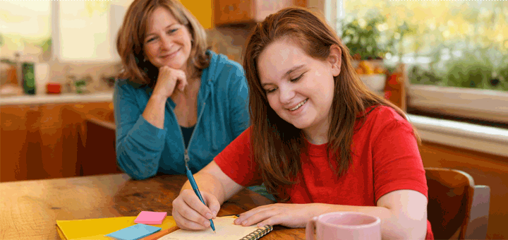Two women smile as one writes in a notebook at a table.
