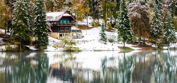 A lodge sits by a calm lake surrounded by snow-dusted trees.