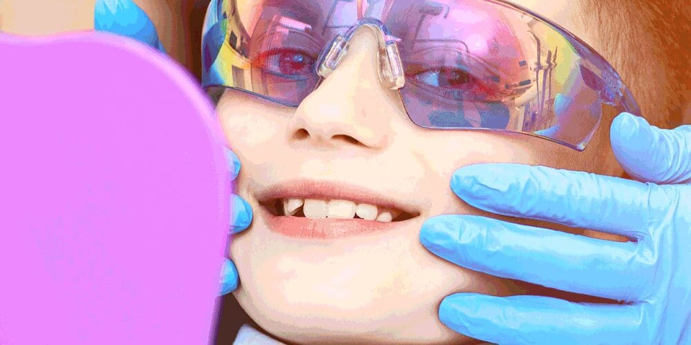 Child wearing sunglasses and blue gloves holds a mirror, with text "Dental visits without the stress" and "Helping children with sensory needs prepare for the dentist.