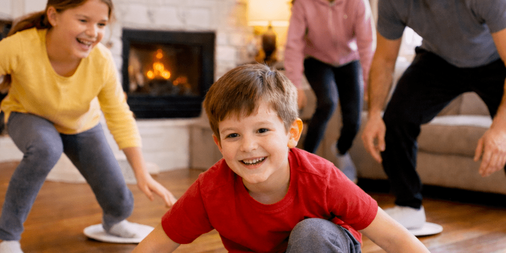 Family playing fitness skating indoors with paper plates on a wooden floor in front of a fireplace.