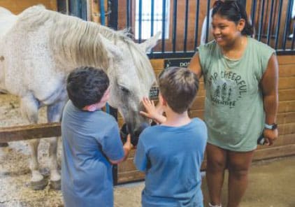 Children pet a white horse in a stall with a woman smiling nearby.