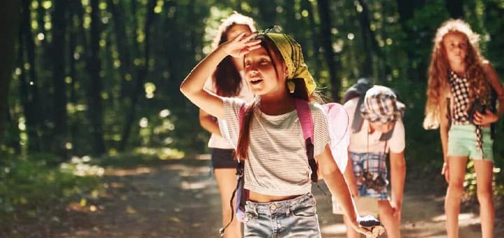 Four girls with backpacks walk down a wooded path on a sunny day.