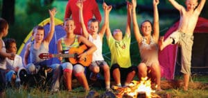 Children with arms raised sit around a campfire, one plays a guitar.