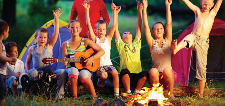 Children with arms raised sit around a campfire, one plays a guitar.