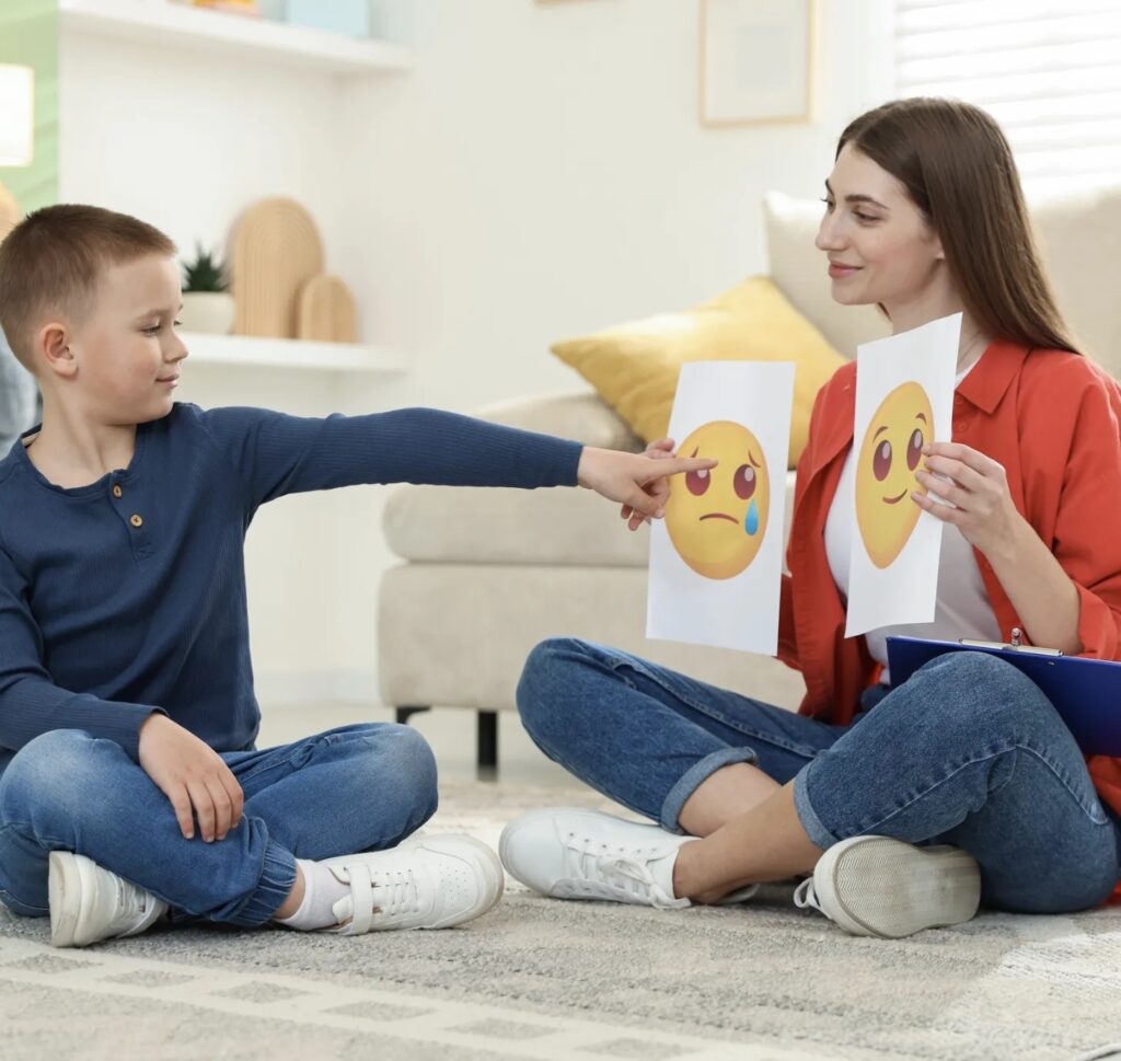 Two people sitting on the floor look at two smiley face drawings on paper.