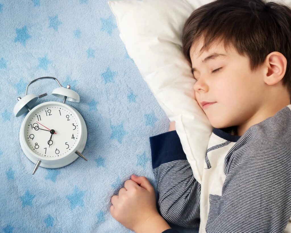 Young boy sleeping with an alarm clock and pillow on a blue surface.