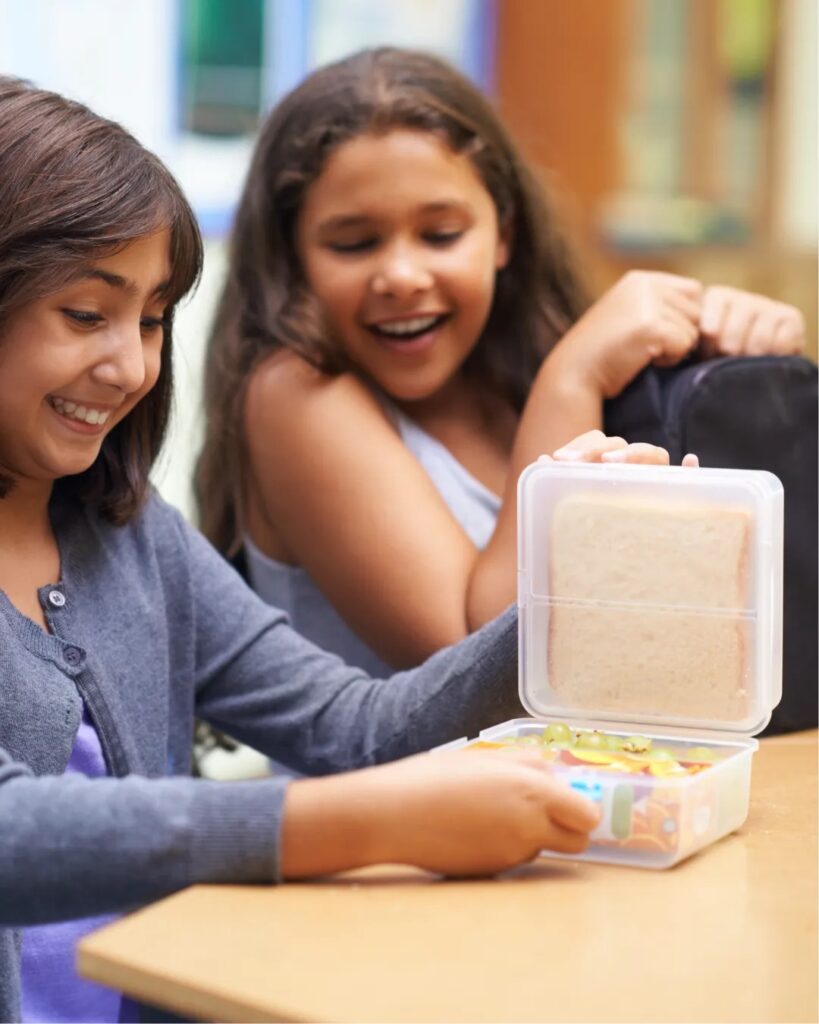 Two girls smiling while eating food from clear plastic containers at a table.